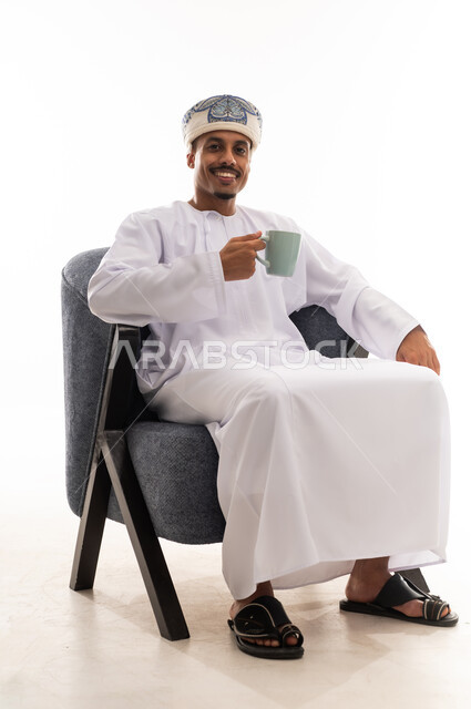 النظر الى الكاميرا و الابتسامة , رجل عربي خليجي عماني يرتدي الثوب التقليدي الابيض العماني يجلس على كرسي رمادي والاستمتاع بالمذاق الشهي , خلفية بيضاء , بورتريه الجسم بالطول الكامل