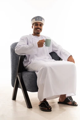 النظر الى الكاميرا و الابتسامة , رجل عربي خليجي عماني يرتدي الثوب التقليدي الابيض العماني يجلس على كرسي رمادي والاستمتاع بالمذاق الشهي , خلفية بيضاء , بورتريه الجسم بالطول الكامل
