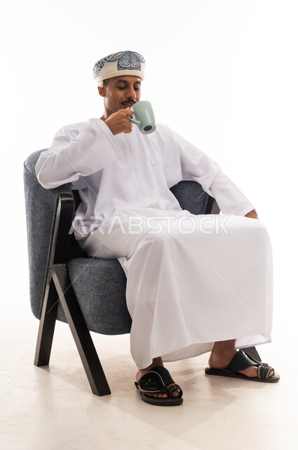 الجلوس في هدوء و احتساء القهوة الساخنة , رجل عربي خليجي عماني يرتدي الدشداشة البيضاء و العمامة , النظر الي الكوب , بورتريه خلفية بيضاء , الجسم بالطول الكامل