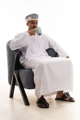 Sitting quietly and sipping hot coffee, an Arab Gulf Omani man wearing a white dishdasha and a turban, looking at the cup, portrait with a white background, full body.