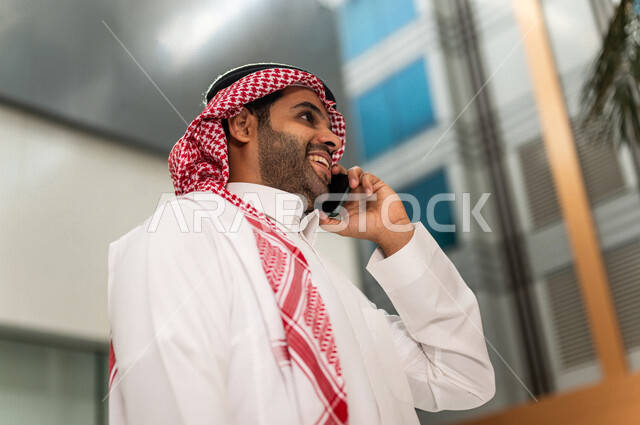 A Saudi Gulf man, making a phone call via a mobile phone inside the tourist hotel, using modern technical equipment, a working session, the concept of business management, making business deals, working remotely, e-marketing, a business trip, tourism and