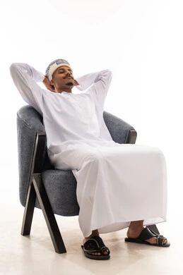 Sitting on the comfortable gray chair and inhaling the gentle breeze and beautiful scent, an Arab Omani Gulf man wearing a white dishdasha and a turban feels relaxed and at ease, portrait with a white background, full-body.