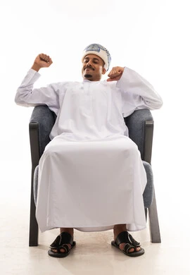 Stretching on the comfortable gray chair, an Arab Gulf Omani man wearing a white dishdasha and a turban clenches his fists and raises them upwards as a sign of stretching and feeling relaxed and rejuvenated, portrait with a white background, full body.