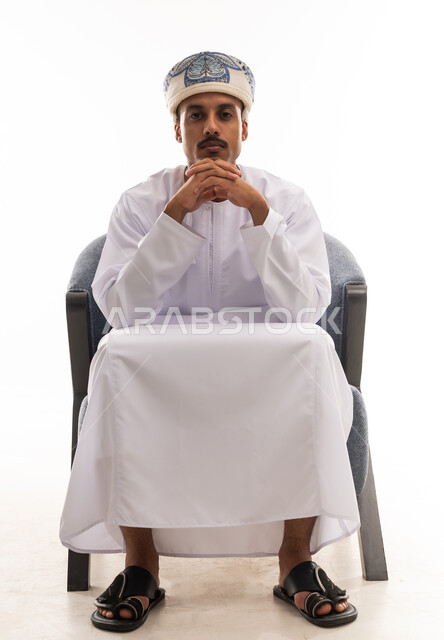 رجل عربي خليجي عماني يرتدي الدشداشة البيضاء و العمامة يجلس على كرسي رمادي , تشبيك اصابع اليدين مع بعضهم اشارة الي التفكير العميق , بورتريه خلفية بيضاء , الجسم بالطول الكامل