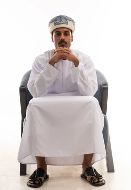 رجل عربي خليجي عماني يرتدي الدشداشة البيضاء و العمامة يجلس على كرسي رمادي , تشبيك اصابع اليدين مع بعضهم اشارة الي التفكير العميق , بورتريه خلفية بيضاء , الجسم بالطول الكامل
