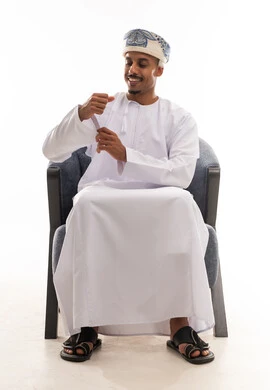 تعديل الثوب الابيض والشعور بالاناقة و الثقة , رجل عربي خليجي عماني يرتدي دشداشة بيضاء و العمامة يجلس على كرسي رمادي اللون , ايماءات الفرحة , الجسم بالطول الكامل , بورتريه خلفية بيضاء