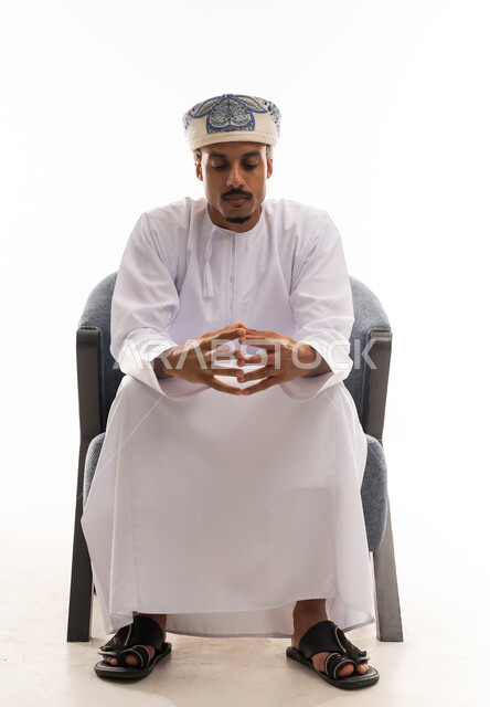 Looking at the fingers of the hands is a sign of confusion and decision-making. An Arab Gulf Omani man wearing a white dishdasha and a turban sits on a gray chair, thinking deeply. The background is white, and it is a full-body portrait.