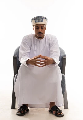 Looking at the fingers of the hands is a sign of confusion and decision-making. An Arab Gulf Omani man wearing a white dishdasha and a turban sits on a gray chair, thinking deeply. The background is white, and it is a full-body portrait.