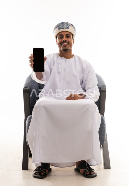 He raised the mobile phone in front of the camera, displaying a blank black screen. An Arab Gulf Omani man wearing a white dishdasha and turban is sitting happily, with gestures of joy and delight, in a portrait with a white background, full-body.