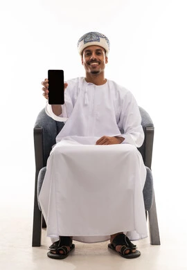 رفع الهاتف المحمول امام الكاميرا , عرض شاشة سوداء فارغة , رجل عربي خليجي عماني يرتدي الدشداشة البيضاء و العمامة , الجلوس بسعادة , ايماءات البهجة و السرور , بورتريه خلفية بيضاء , الجسم بالطول الكامل