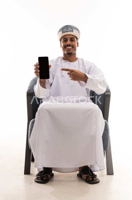 Pointing with the index finger at a blank black mobile phone screen, an Arab Gulf Omani man wearing a white dishdasha and a turban sits on a gray chair, showing gestures of happiness and joy, portrait with a white background, full body.