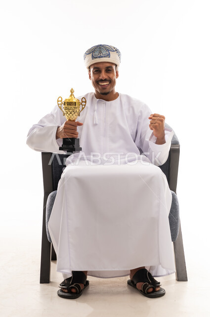 الابتسامة الى الكاميرا اشارة على السعادة للفوز بالكأس الذهبية , رجل عربي خليجي عماني يرتدي الدشداشة البيضاء و العمامة , قفل قبضة اليد علامة على البهجة بتحقيق الفوز و رفع الكأس باليد الاخرى , خلفية بيضاء , بورتريه الجسم بالطول الكامل