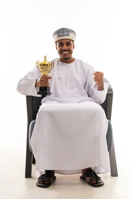 الابتسامة الى الكاميرا اشارة على السعادة للفوز بالكأس الذهبية , رجل عربي خليجي عماني يرتدي الدشداشة البيضاء و العمامة , قفل قبضة اليد علامة على البهجة بتحقيق الفوز و رفع الكأس باليد الاخرى , خلفية بيضاء , بورتريه الجسم بالطول الكامل