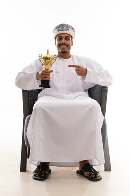 رفع كأس الجائزة الكبرى للاعلي , رجل عربي خليجي عماني يرتدي الثوب الابيض التقليدي في سلطنة عمان يشير باصبع السبابة الى الكأس الذهبية , بورتريه الجسم بالطول الكامل , خلفية بيضاء