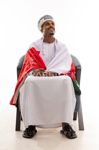 النظر للاعلى الى شئ ما , رجل عربي خليجي عماني يرتدي الدشداشة البيضاء و عمامة , الجلوس على الكرسي و وضع العلم العماني على الاكتاف , بورتريه الجسم بالطول الكامل , خلفية بيضاء