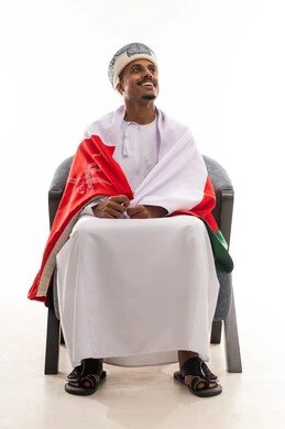 النظر للاعلى الى شئ ما , رجل عربي خليجي عماني يرتدي الدشداشة البيضاء و عمامة , الجلوس على الكرسي و وضع العلم العماني على الاكتاف , بورتريه الجسم بالطول الكامل , خلفية بيضاء