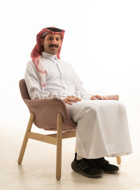 الأناقة والوسامة والاهتمام بالمظهر الخارجي، بورتريه لرجل عربي خليجي سعودي يرتدي الزي التقليدي و الشماغ يجلس على كرسي ينظر للكاميرا بتعابير السعادة و السرور ، خلفية بيضاء