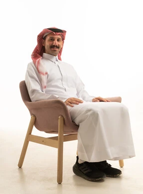 Elegance, handsomeness, and attention to appearance; a portrait of a Saudi Arabian Gulf man wearing traditional attire and a ghutrah, sitting on a chair looking at the camera with expressions of happiness and joy, with a white background.