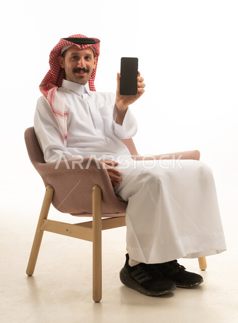 رجل سعودي يرتدي الزي التقليدي يحمل هاتف ذكي بخلفية بيضاء