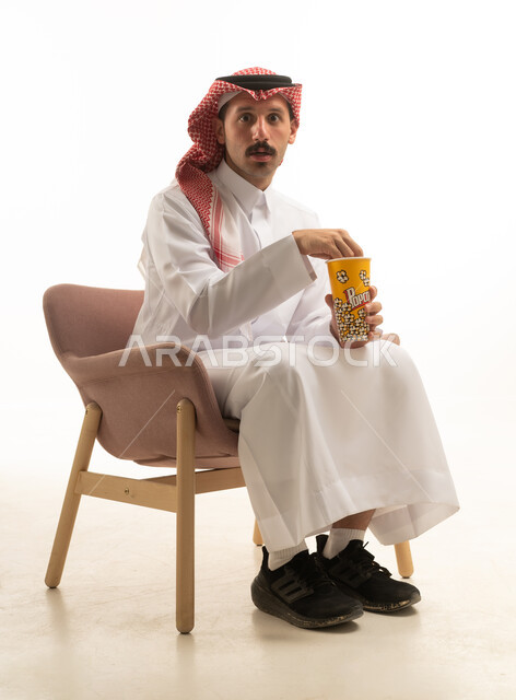 رجل سعودي مصدوم يجلس مع علبة فشار خلفية بيضاء