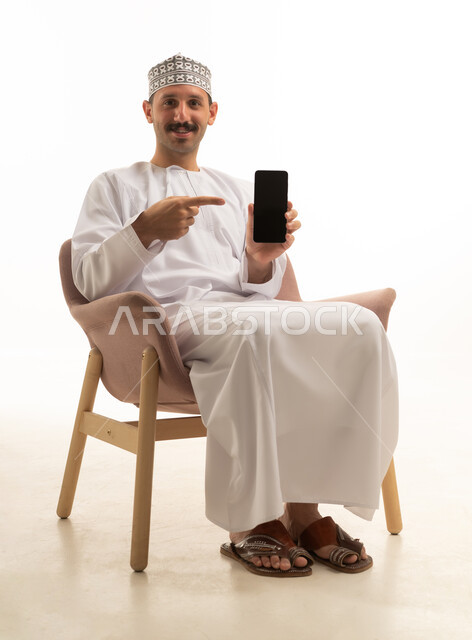 رجل عماني عربي خليجي يرتدي الكمه والدشداشه يمسك الهاتف المحمول يجلس على كرسي بايماءات وحركات متنوعه, خلفيه بيضاء