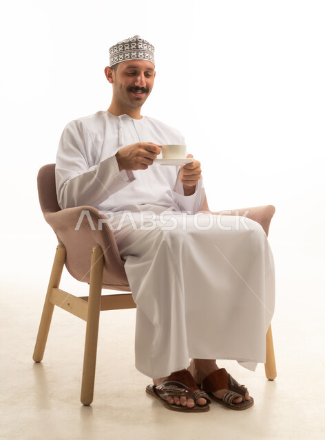 رجل عماني عربي يرتدي الدشداشه والكمه يجلس على كرسي ويشرب فنجان قهوه, الاستمتاع بالمشروب الساخن, خلفيه بيضاء