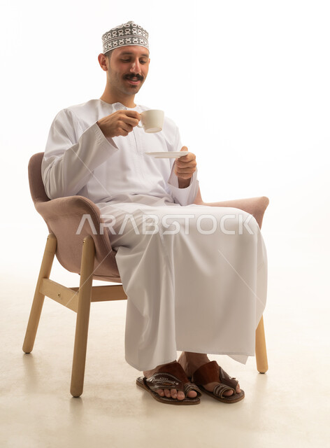 رجل عماني عربي يرتدي الدشداشه والكمه يجلس على كرسي ويشرب فنجان قهوه, الاستمتاع بالمشروب الساخن, خلفيه بيضاء
