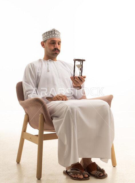 رجل عماني يجلس مع ساعة رملية بخلفية بيضاء