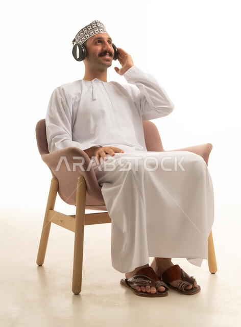 رجل عربي خليجي عماني يرتدي الدشداشه والكمه يجلس على كرسي يرتدي سماعة الراس, التسليه والترفيه, خلفيه بيضاء