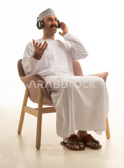 رجل عربي خليجي عماني يرتدي الدشداشه والكمه يجلس على كرسي يرتدي سماعة الراس, التسليه والترفيه, خلفيه بيضاء