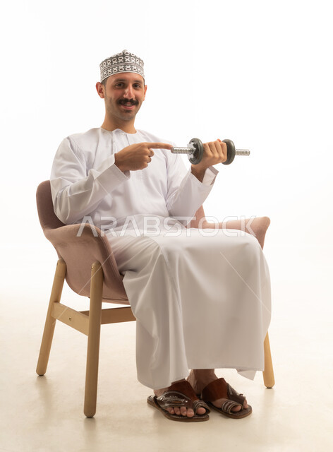 رجل عربي خليجي عماني يرتدي الدشداشه والكمه يجلس على كرسي ويمسك بيده الدمبلز, نشاط بدني ممتع, بناء العضلات والصحه واللياقه الجسديه, التسليه والترفيه, خلفيه بيضاء