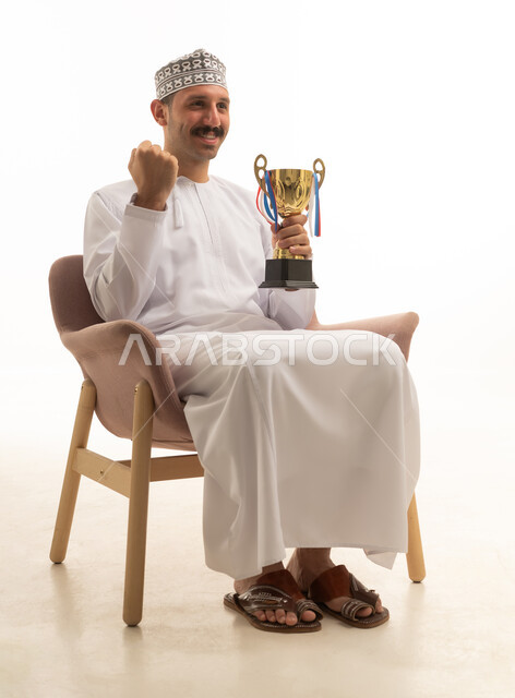 بورتريه لرجل عماني عربي خليجي يرتدي الدشداشه والكمه يجلس على كرسي يمسك بيده كاس الفوز, تشجيع الفريق الرياضي, خلفيه بيضاء