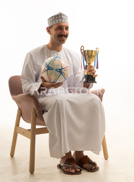 بورتريه لرجل عماني عربي خليجي يرتدي الدشداشه والكمه يجلس على كرسي يمسك بيده كرة قدم وكاس الفوز, تشجيع الفريق الرياضي, خلفيه بيضاء
