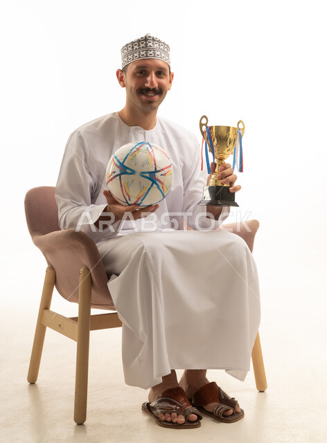 A portrait of an Omani Arab Gulf man wearing a dishdasha and kumma sitting on a chair holding a football and a trophy, cheering for the sports team, with a white background.