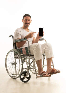 A portrait of a man with disabilities, an Arab Saudi Gulf, wearing pants and a shirt, sitting in a wheelchair, pointing to a blank black screen through a mobile phone, with a white background.
