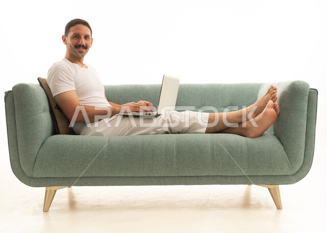 رجل سعودي يستخدم اللابتوب على أريكة حديثة بخلفية بيضاء