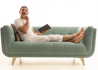 A Saudi Arabian Gulf Arab man wearing a shirt and white pants is sitting on a comfortable green sofa reading a book, with a white background.