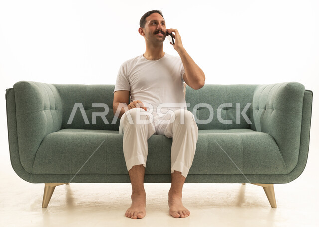 رجل عربي سعودي خليجي يرتدي الفنيلا والسروال الابيض يجلس على اريكه خضراء مريحه, استخدام الهاتف المحمول, خلفيه بيضاء