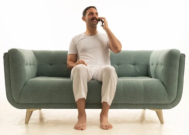 A Saudi Arabian Gulf Arab man wearing a t-shirt and white pants is sitting on a comfortable green sofa, using a mobile phone, with a white background.