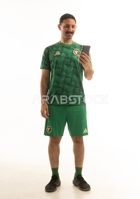 لاعب كرة قدم سعودي يرتدي الزي الرياضي الاخضر يمسك بيده الهاتف المحمول, ايماءات وحركات متنوعه, كامل الجسم, خلفيه بيضاء