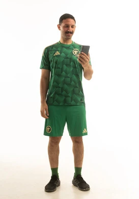 لاعب كرة قدم سعودي يرتدي الزي الرياضي الاخضر يمسك بيده الهاتف المحمول, ايماءات وحركات متنوعه, كامل الجسم, خلفيه بيضاء