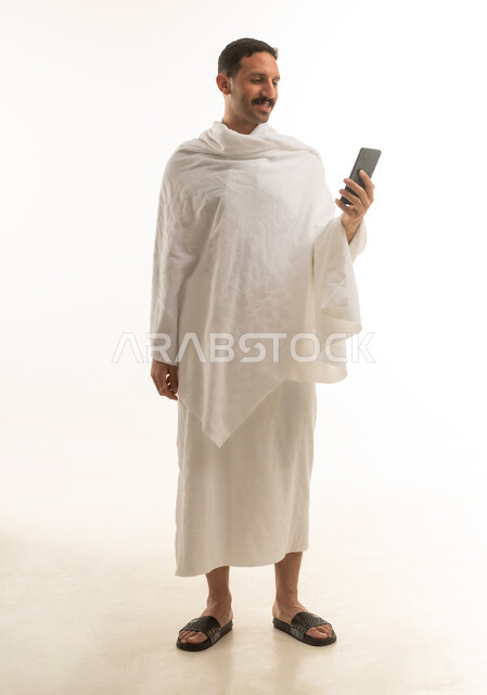 بورتريه لرجل عربي سعودي خليجي يرتدي ملابس الاحرام البيضاء ويمسك بيده الهاتف المحمول, اداء مناسك الحج والعمره, العباده والتقرب الى الله, خلفيه بيضاء , الجسم بالطول الكامل
