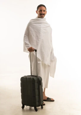 A portrait of an Arab Saudi Gulf man wearing white Ihram clothing, standing with a travel bag in hand, performing the rituals of Hajj and Umrah, worship and drawing closer to God, with a white background, full-body.