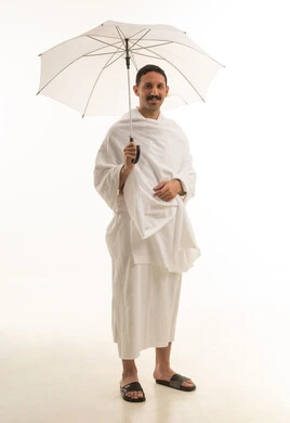 A portrait of an Arab Saudi Gulf man wearing white Ihram clothing, standing and holding a white umbrella, performing the rituals of Hajj and Umrah, worship and drawing closer to God, with a white background, full-body.