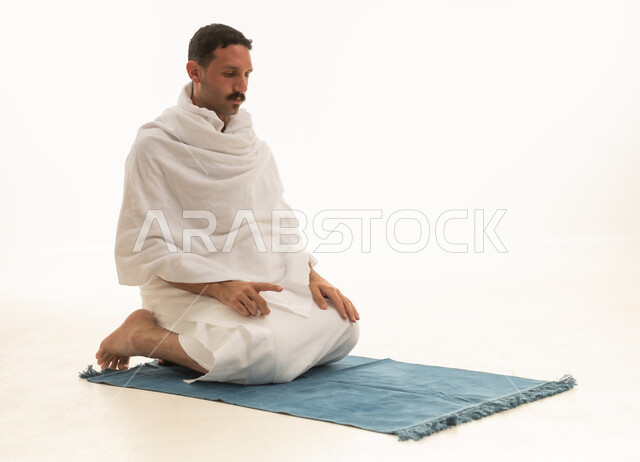 A portrait of an Arab Saudi Gulf man wearing white Ihram clothing sitting on a prayer rug and praying, performing the rituals of Hajj and Umrah, worship and drawing closer to God, with a white background.