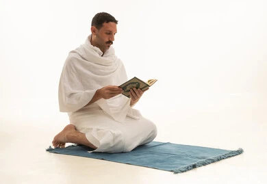 التقرب الى رب العالمين في شهر رمضان المبارك، قراءة كتاب الله تعالى, إيماءات الخشوع و الإجتهاد في العباده, بورتريه  لرجل عربي سعودي خليجي يرتدي ملابس الاحرام يمسك بيده القرآن الكريم يجلس على سجادة الصلاه، خلفية بيضاء