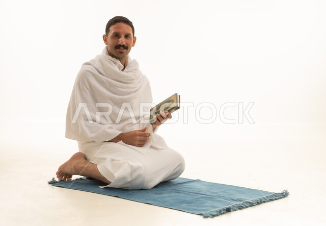 قراءة كتاب الله تعالى, إيماءات الخشوع و الإجتهاد في العباده, بورتريه  لرجل عربي سعودي خليجي يرتدي ملابس الاحرام يمسك بيده القرآن الكريم يجلس على سجادة الصلاه, التقرب الى رب العالمين في شهر رمضان المبارك، خلفية بيضاء