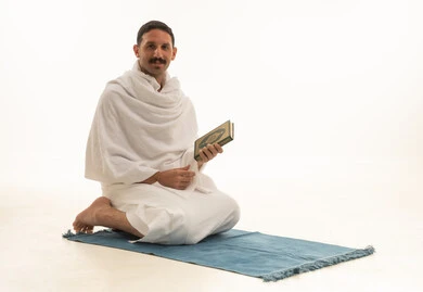 قراءة كتاب الله تعالى, إيماءات الخشوع و الإجتهاد في العباده, بورتريه  لرجل عربي سعودي خليجي يرتدي ملابس الاحرام يمسك بيده القرآن الكريم يجلس على سجادة الصلاه, التقرب الى رب العالمين في شهر رمضان المبارك، خلفية بيضاء