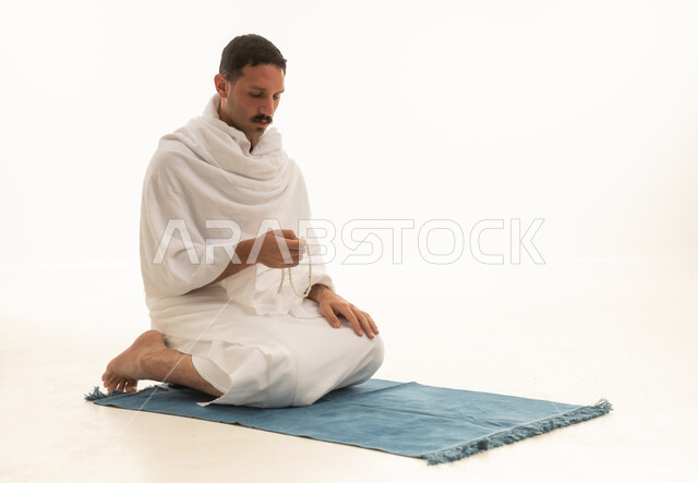 الإجتهاد في العباده والتقرب الى الله في شهر رمضان المبارك, مفهوم التسبيح والتهليل، بورتريه لرجل عربي خليجي سعودي يرتدي ملابس الاحرام يجلس على سجادة الصلاة يمسك بيده السبحه, الإكثار من ذكر الله تعالى من خلال الإستغفار والدعاء, خلفيه بيضاء