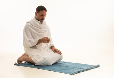 الإجتهاد في العباده والتقرب الى الله في شهر رمضان المبارك, مفهوم التسبيح والتهليل، بورتريه لرجل عربي خليجي سعودي يرتدي ملابس الاحرام يجلس على سجادة الصلاة يمسك بيده السبحه, الإكثار من ذكر الله تعالى من خلال الإستغفار والدعاء, خلفيه بيضاء